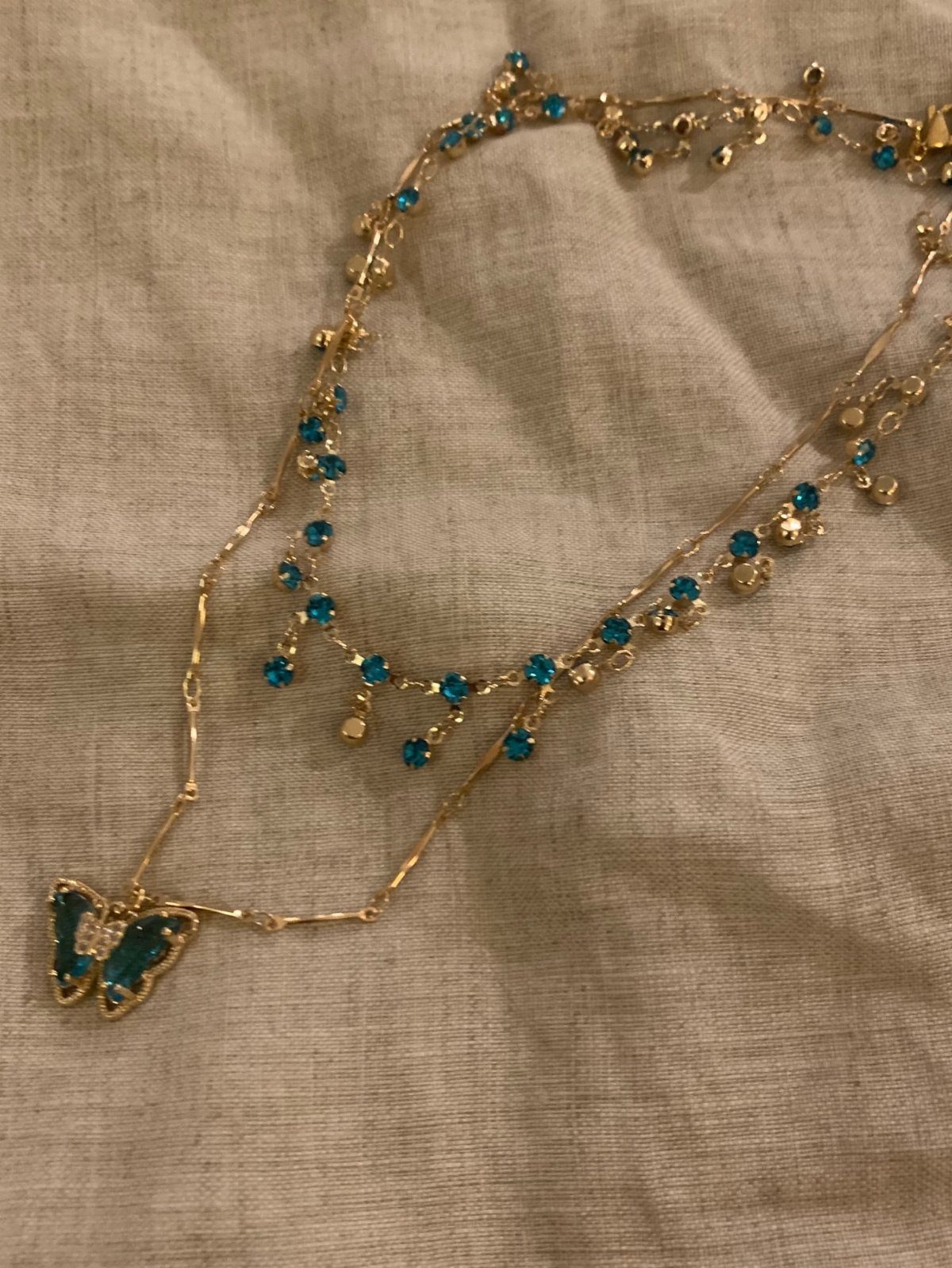 13 Going on 30 Outfits: 6 Ideas to Recreate the Iconic Style 2 Gold necklace with blue beads and a butterfly pendant on beige fabric.