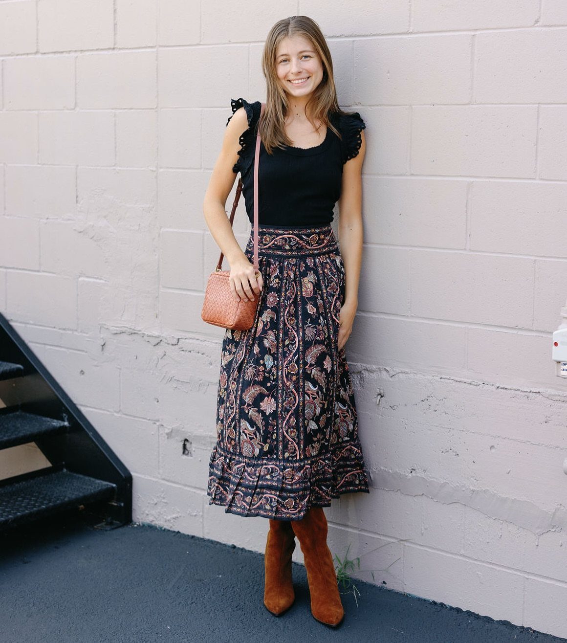 Woman in black top and patterned bohemian skirt posing against a purple wall