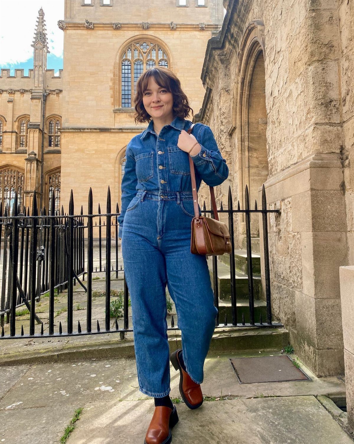 Woman in a blue denim jumpsuit and brown boots standing by a stone building.