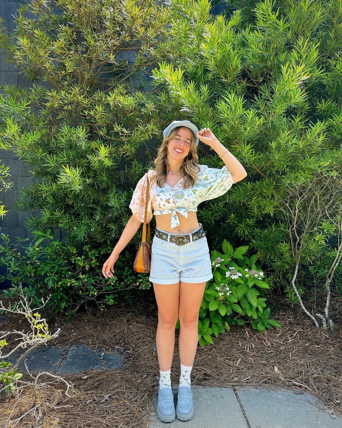 Woman wearing floral crop top, denim shorts, bucket hat, and grey sneakers outdoors.