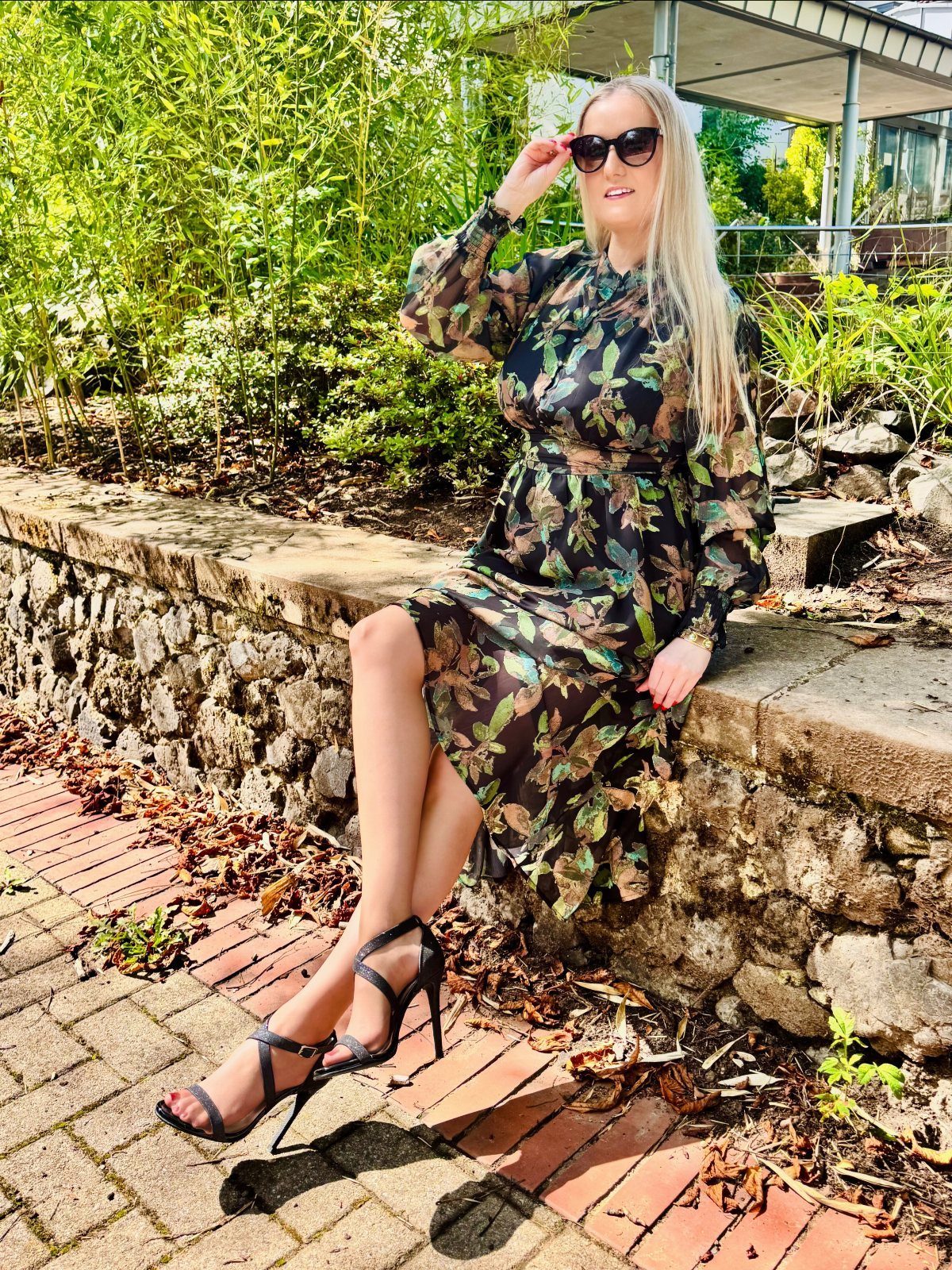 Woman in floral midi dress and black heels sitting on a stone wall.