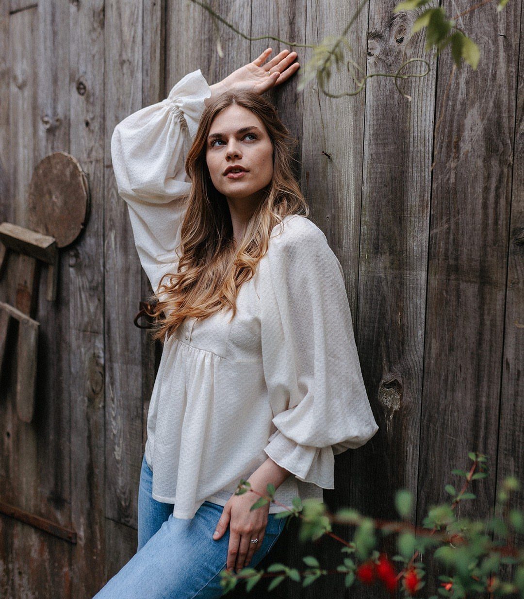 Woman in white puff-sleeve blouse and blue jeans leaning against a wooden fence.