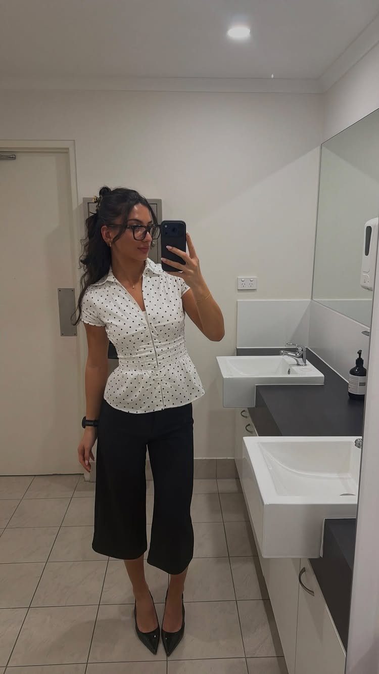Woman in a white polka dot shirt and black trousers posing in a bathroom.