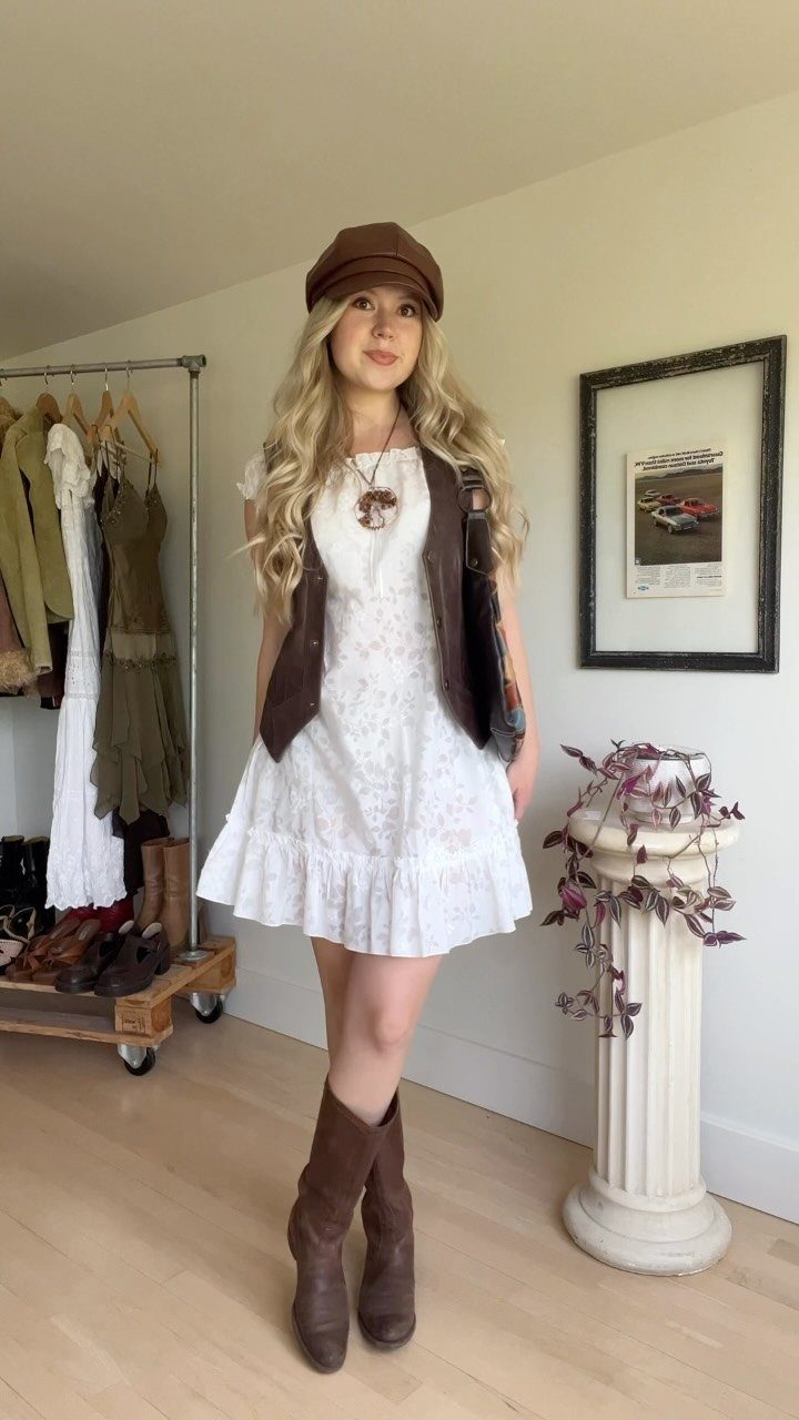 Woman wearing a white floral babydoll dress with a brown vest and cap.