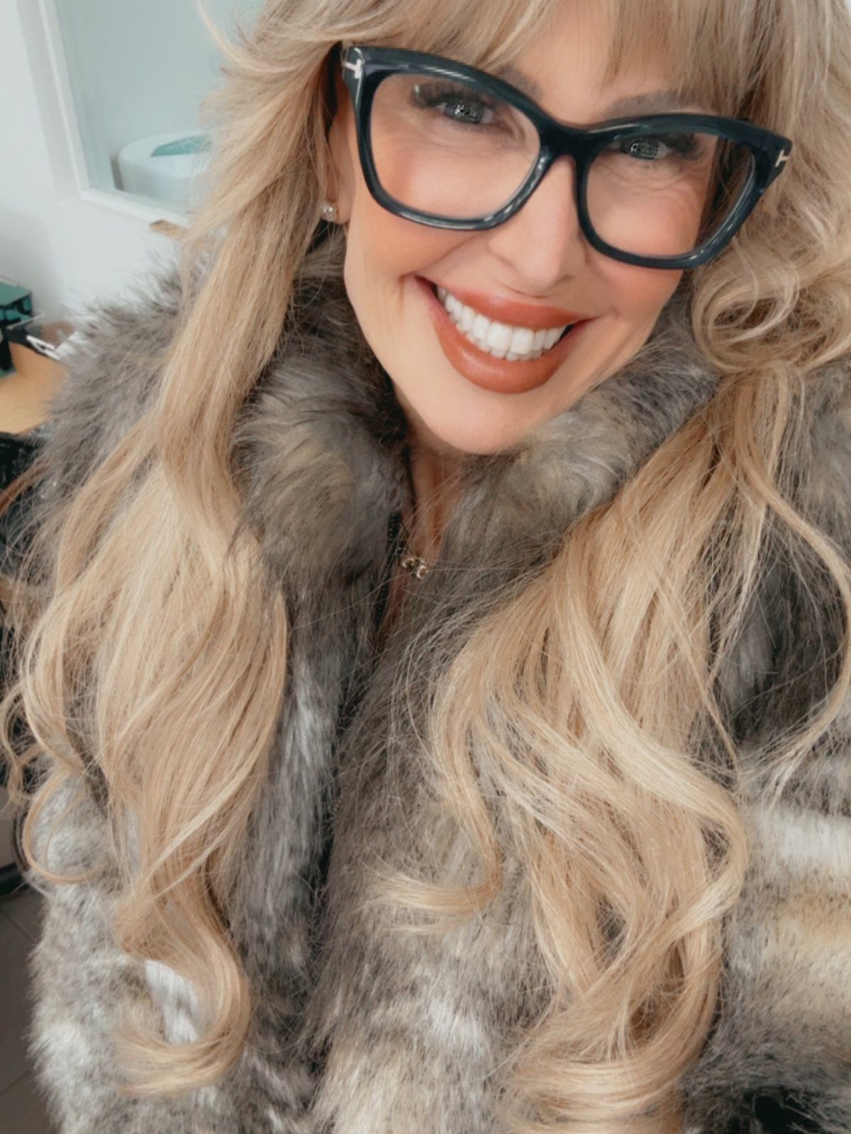 Woman with blonde hair and glasses wearing a grey faux fur coat.