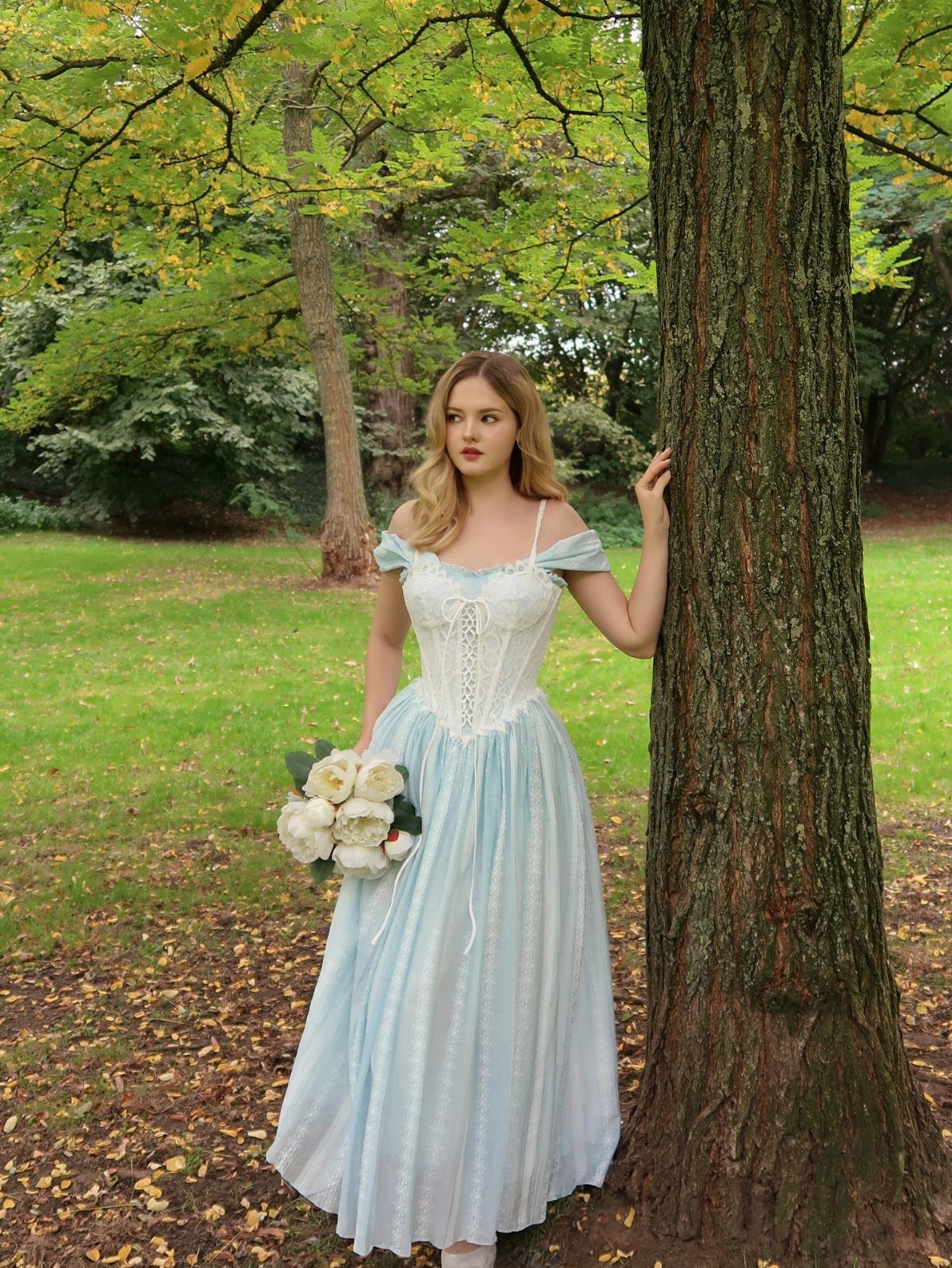 Woman in light blue gown with white lace bodice holding white roses by a tree.