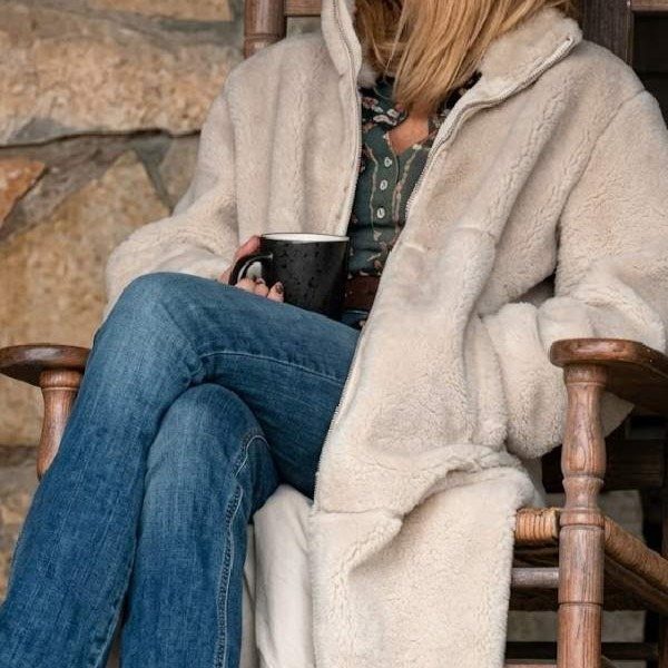 Beth Dutton Outfits: 12 Looks from Yellowstone's Style Icon 7 Person in a cream faux fur coat and blue jeans sitting in a rocking chair.