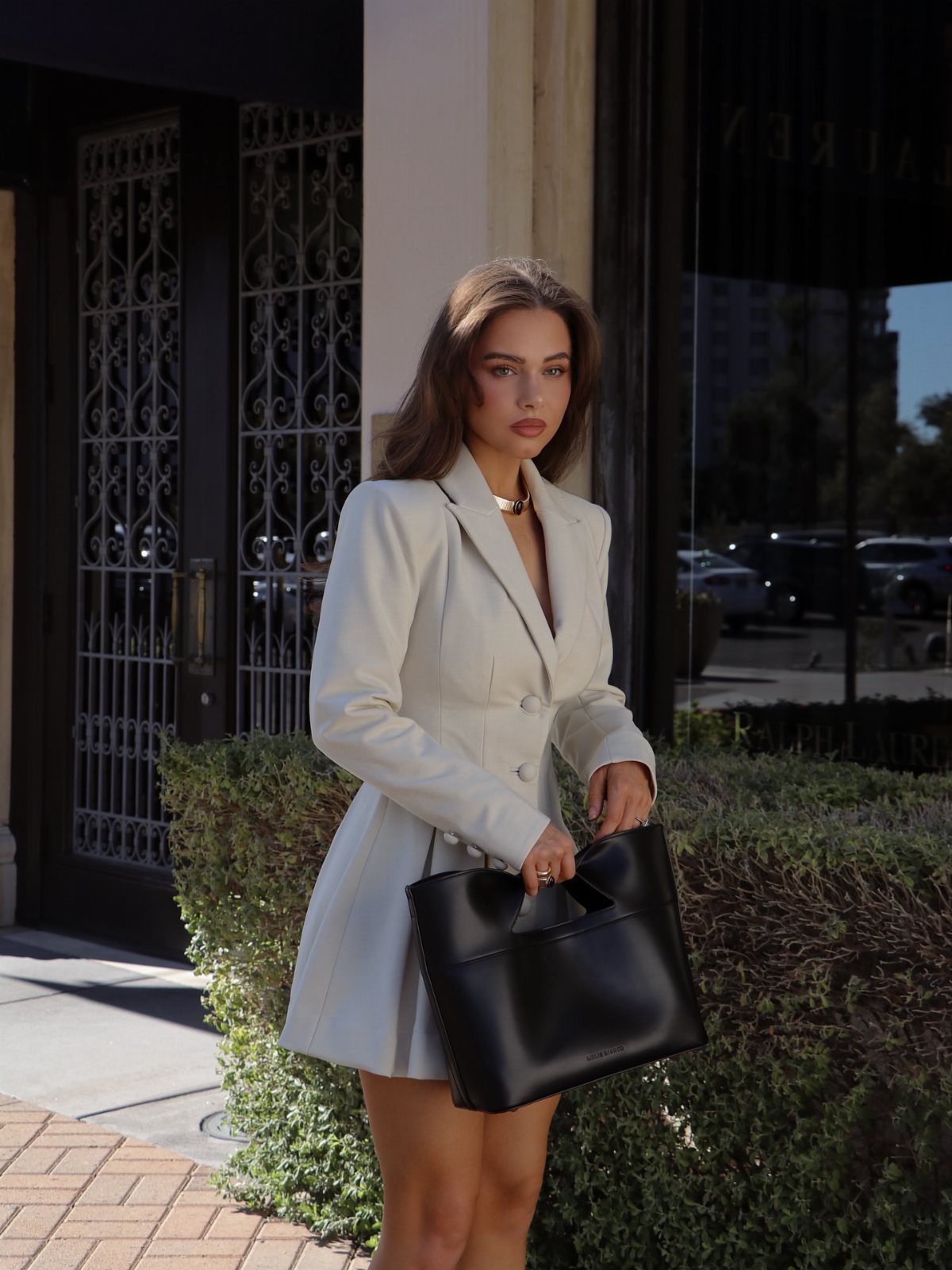 Beth Dutton Outfits: 12 Looks from Yellowstone's Style Icon 3 Woman in a structured off-white blazer dress holding a black leather handbag outdoors.