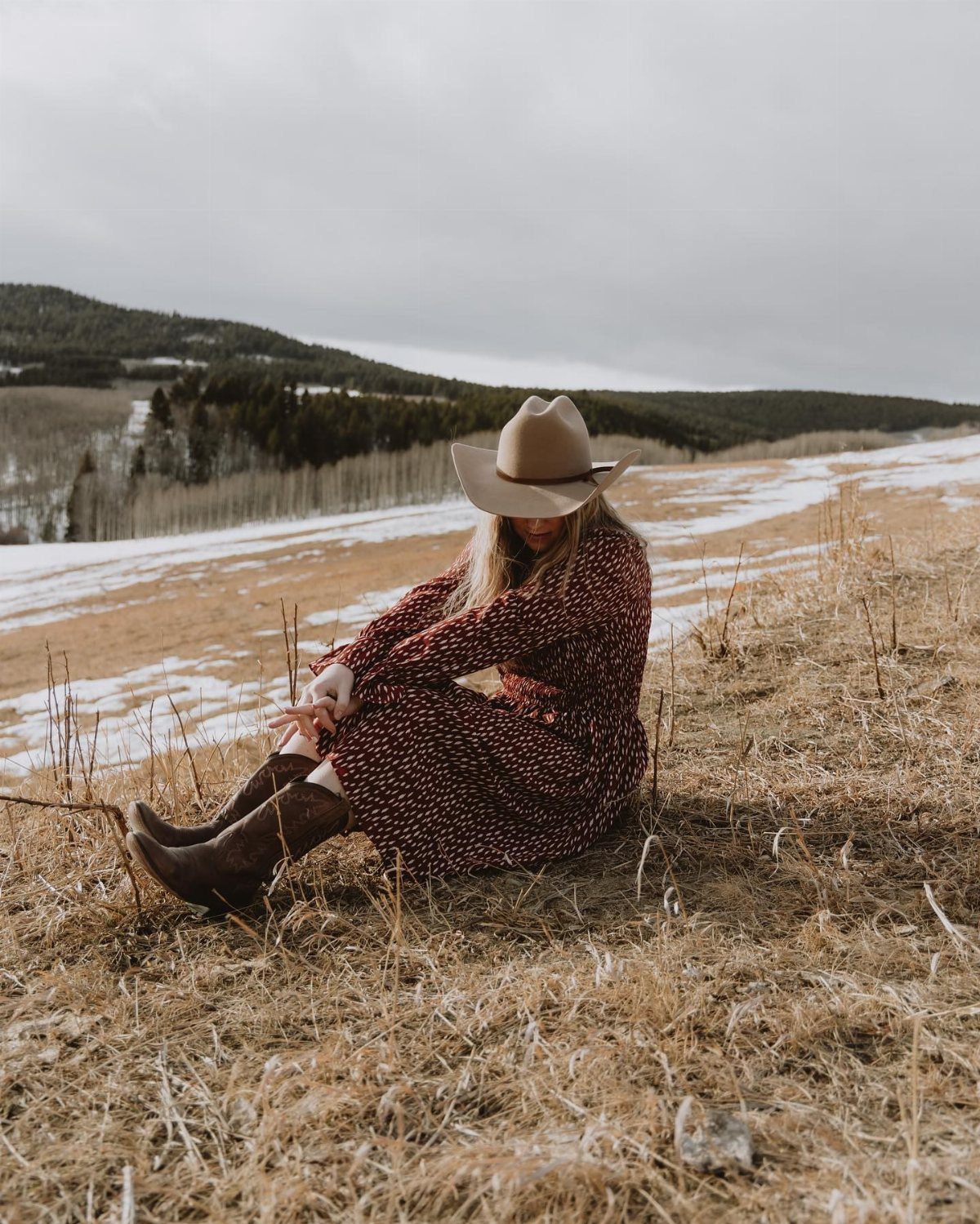 Beth Dutton Outfits: 12 Looks from Yellowstone's Style Icon 2 Woman in a red floral dress and cowboy boots sitting in a winter field.