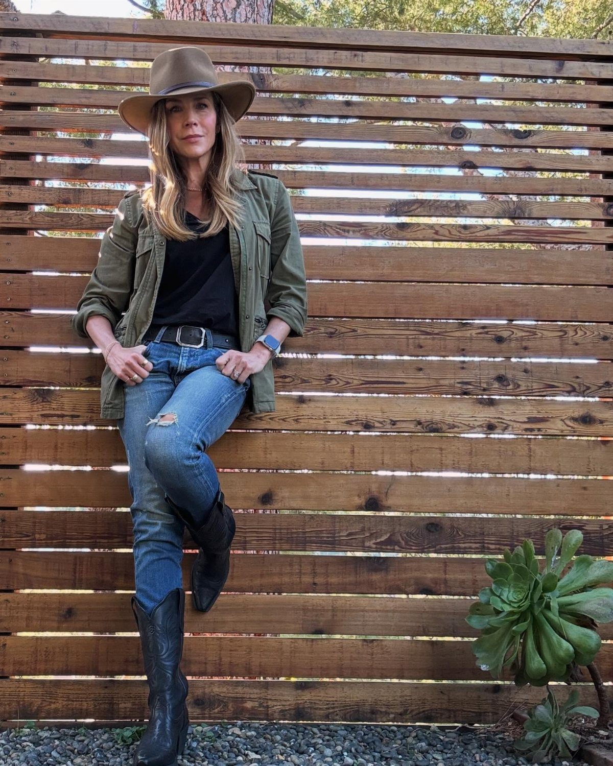 Beth Dutton Outfits: 12 Looks from Yellowstone's Style Icon 11 Woman in olive jacket, cowboy boots, and brown hat leaning on wooden fence