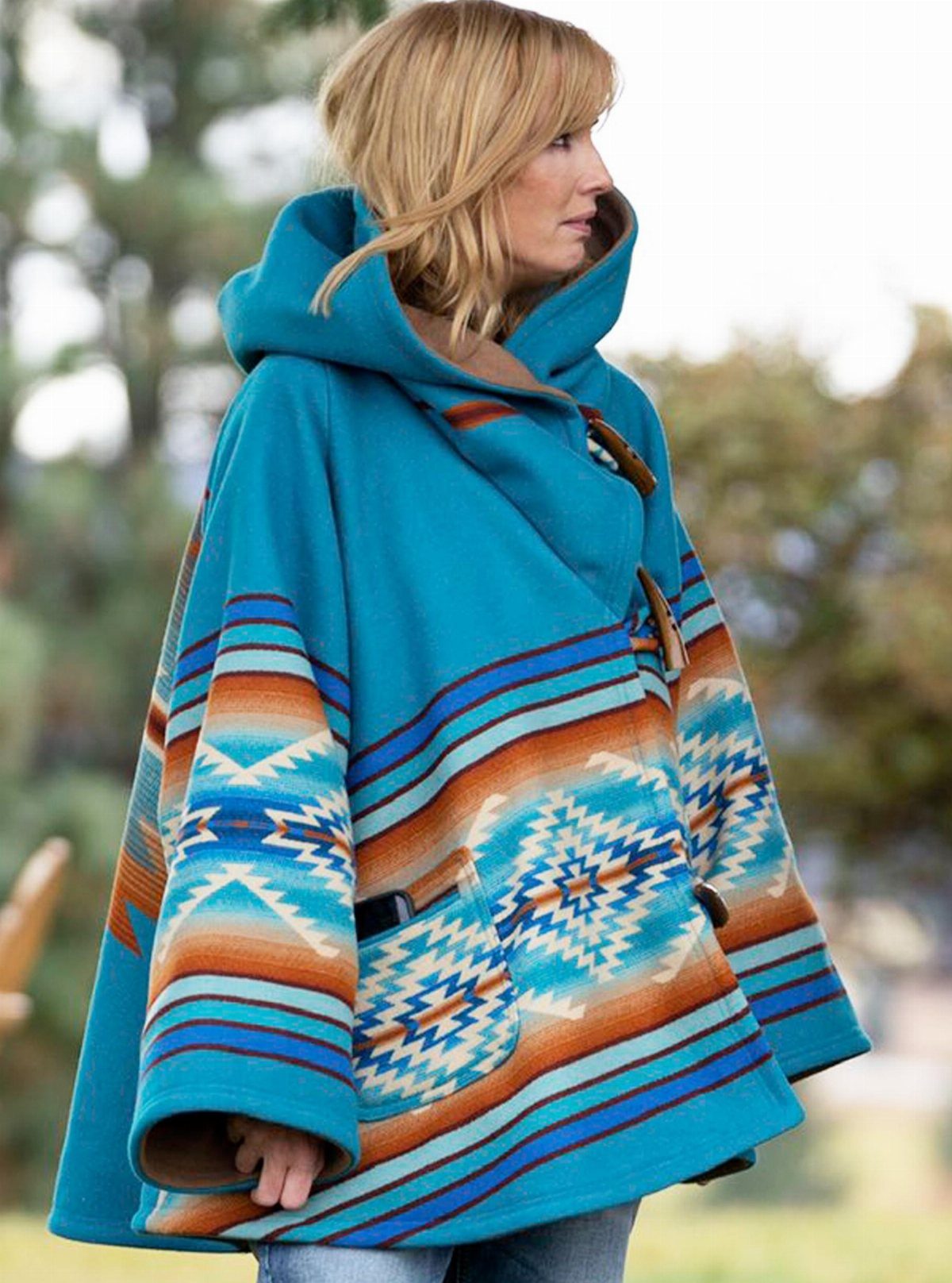Beth Dutton Outfits: 12 Looks from Yellowstone's Style Icon 6 Woman wearing a blue hooded poncho with geometric patterns and blue jeans outdoors.