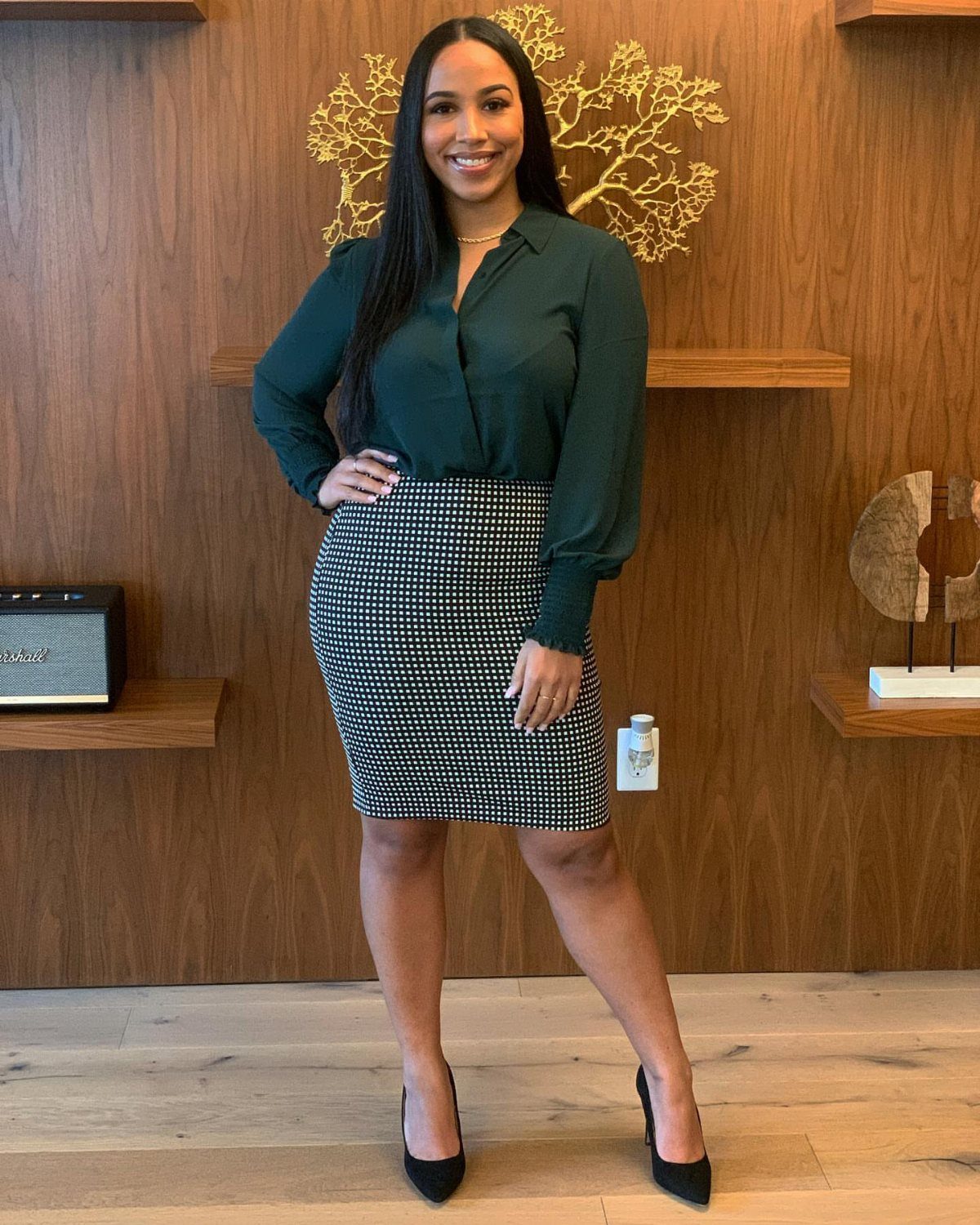 Black woman wearing a green button-down shirt and patterned pencil skirt posing indoors.