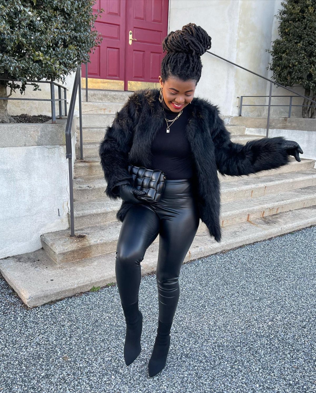 Black woman in a faux-fur coat and leather leggings smiling outdoors by red doors.