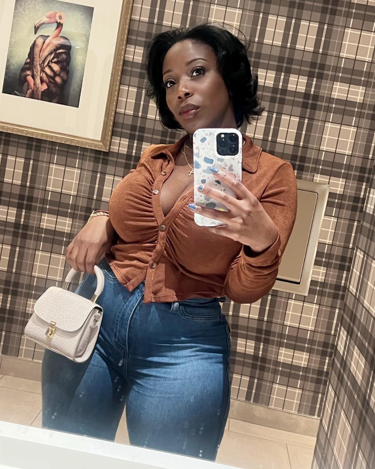 Black woman in a brown form-fitting shirt and blue jeans taking a mirror selfie.