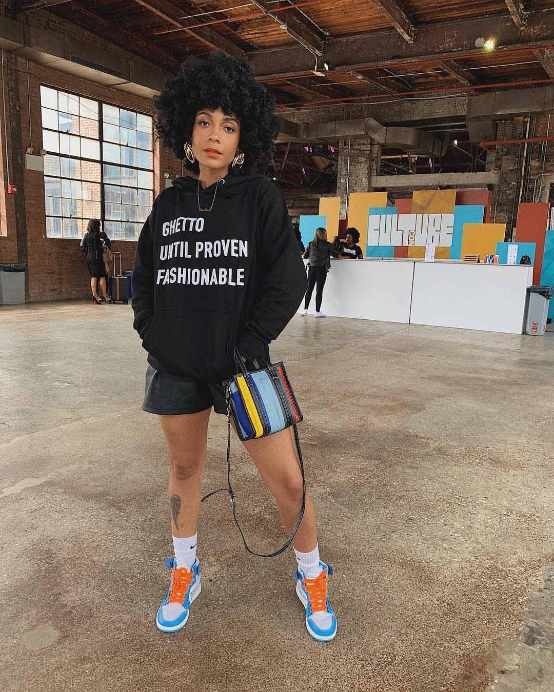 Woman with afro wearing a black graphic hoodie, leather skirt, and colorful sneakers.