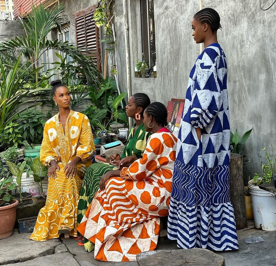 Four Black women wearing colorful African print dresses in an outdoor courtyard setting.