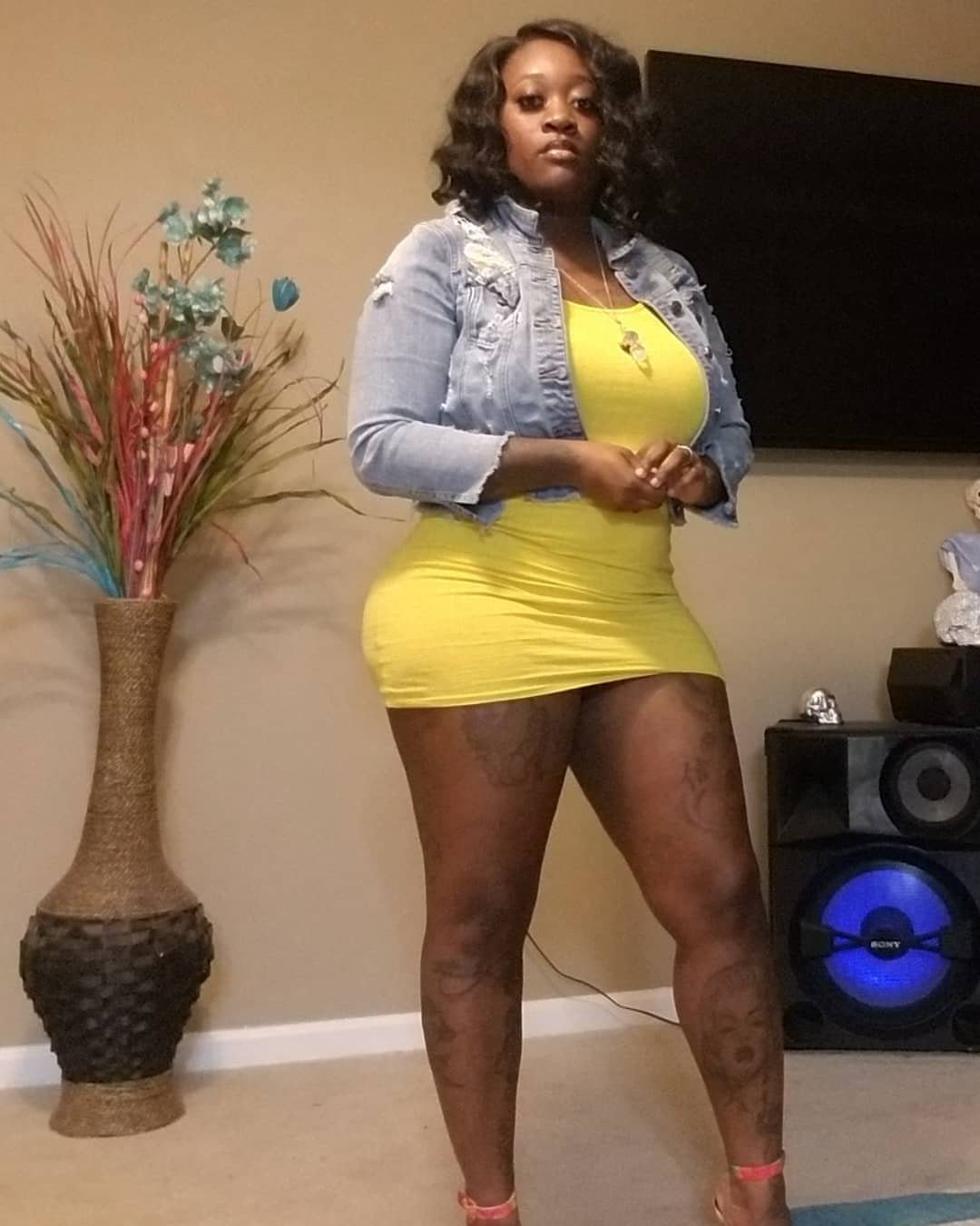 Black woman in a yellow mini dress and denim jacket standing indoors.