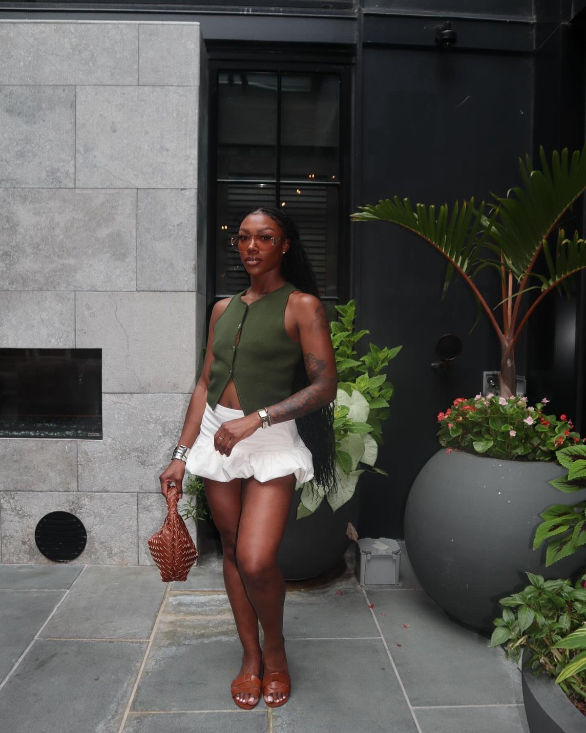 Black woman wearing an olive green vest and white ruffled skirt standing outdoors.