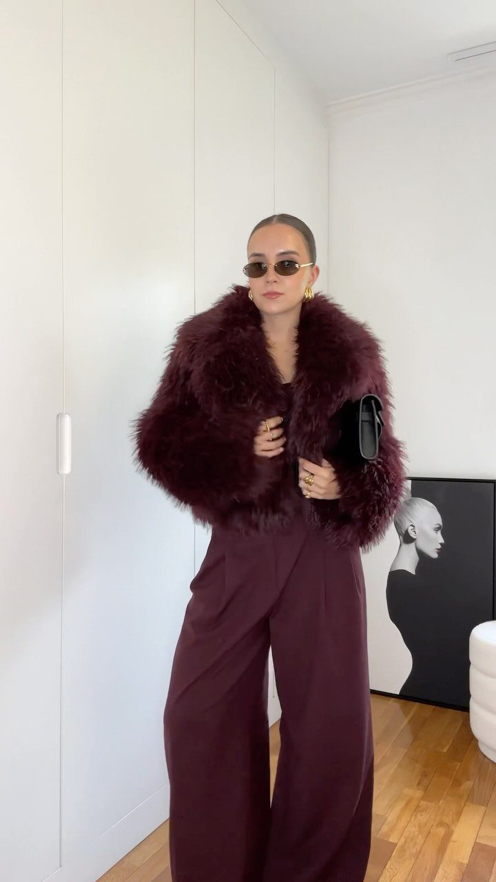 7 Burgundy Outfits for Women to Wear This Season 4 Woman in a burgundy faux-fur jacket and wide-leg burgundy trousers holding a black clutch.