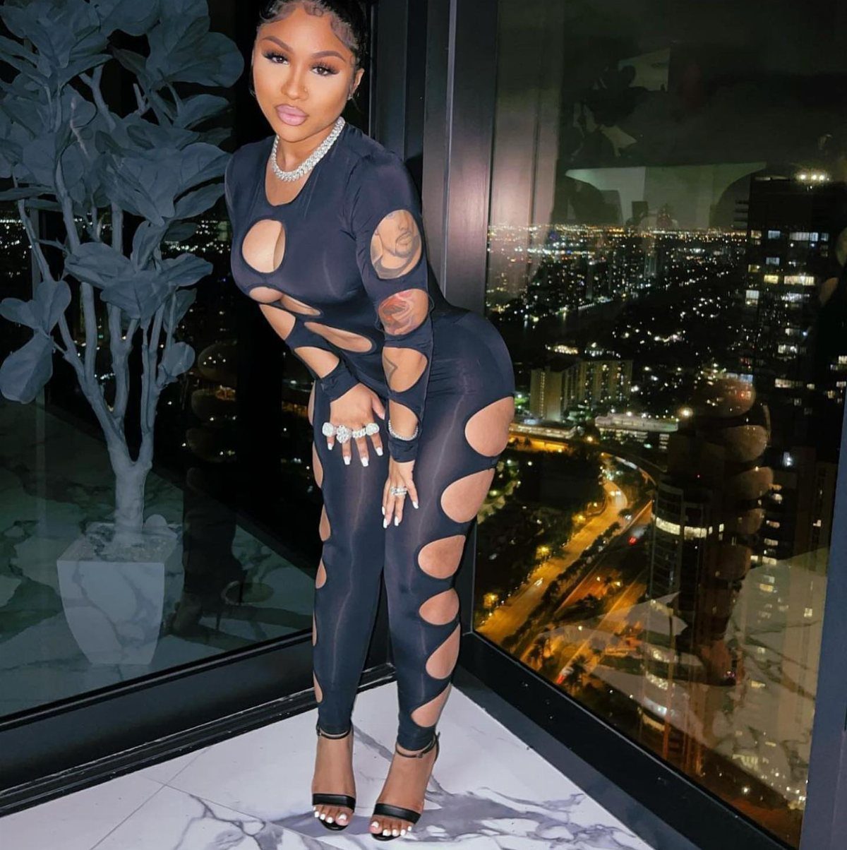 Cardi B Concert Outfits: 9 Bold Looks to Turn Heads 8 Woman in a black cutout bodysuit posing against a nighttime city skyline view.
