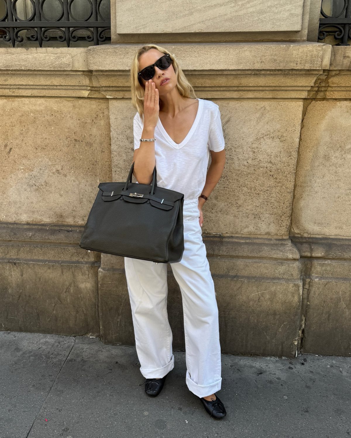 13 Carolyn Bessette Outfits You Can Actually Wear Today 3 Woman in white outfit carrying a large black leather Birkin bag against stone wall.