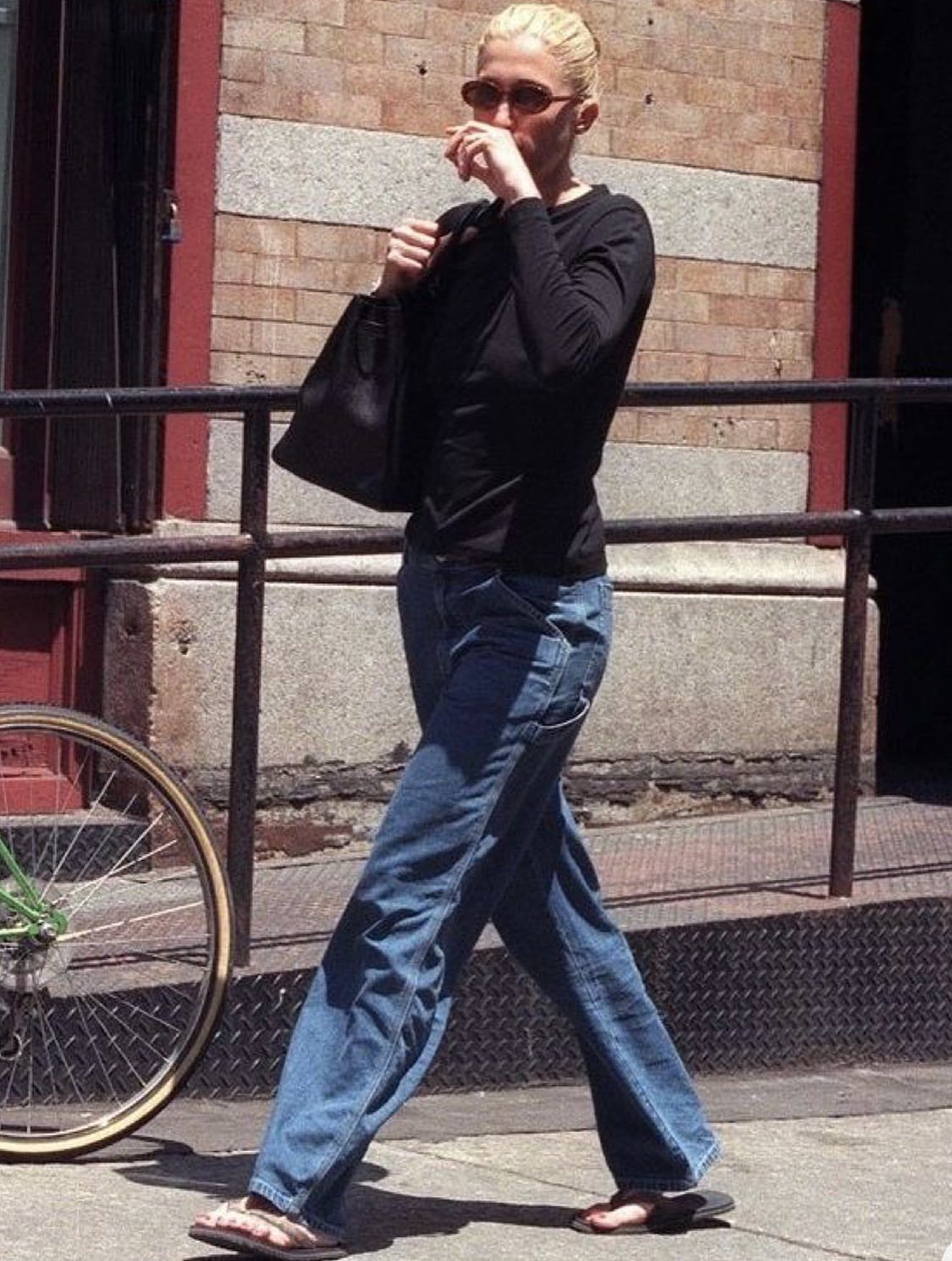 13 Carolyn Bessette Outfits You Can Actually Wear Today 6 Woman in black long-sleeved top and loose blue jeans walking on city sidewalk.