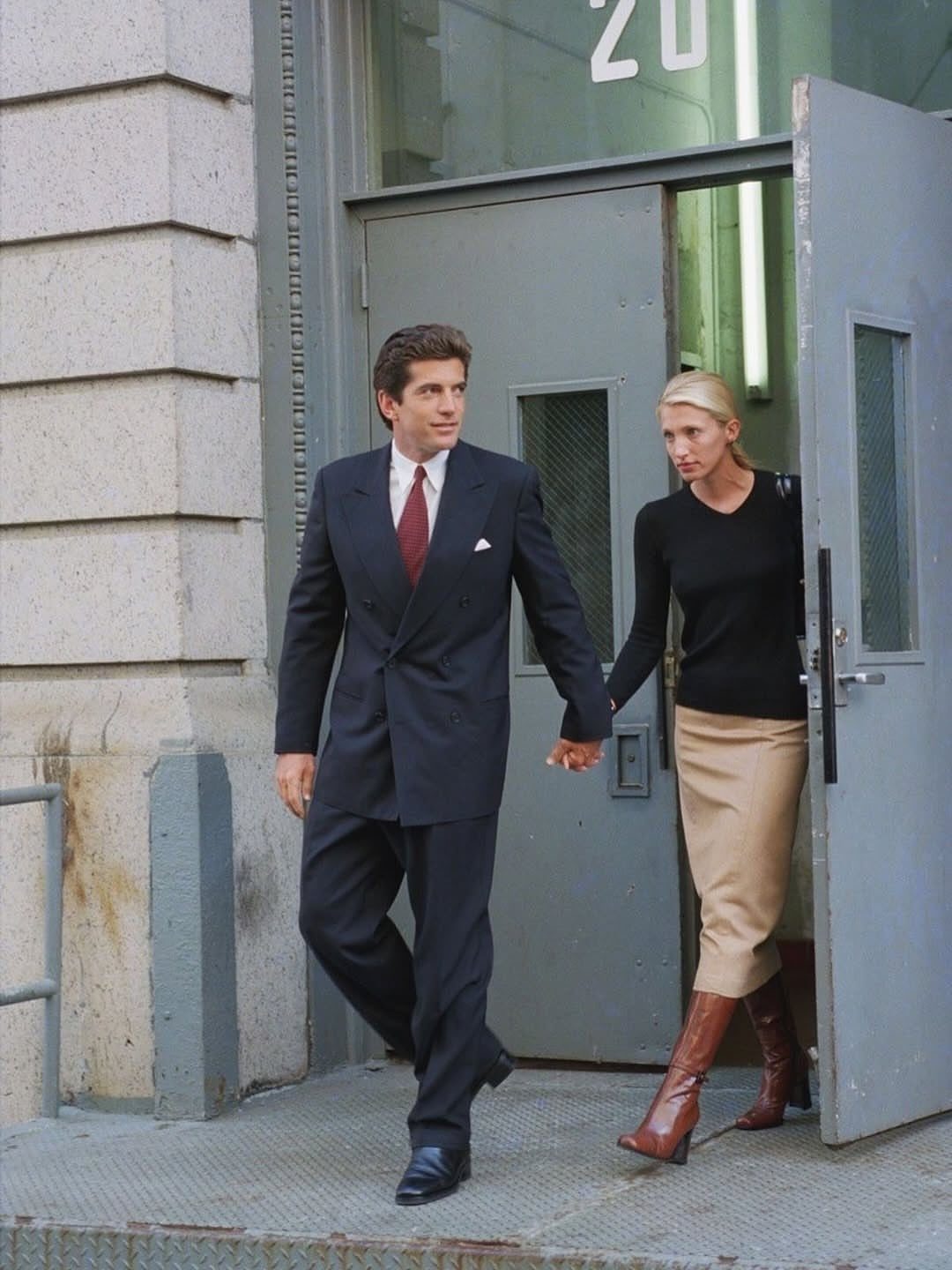 13 Carolyn Bessette Outfits You Can Actually Wear Today 2 Man in dark suit and woman in black top and beige skirt walking together.