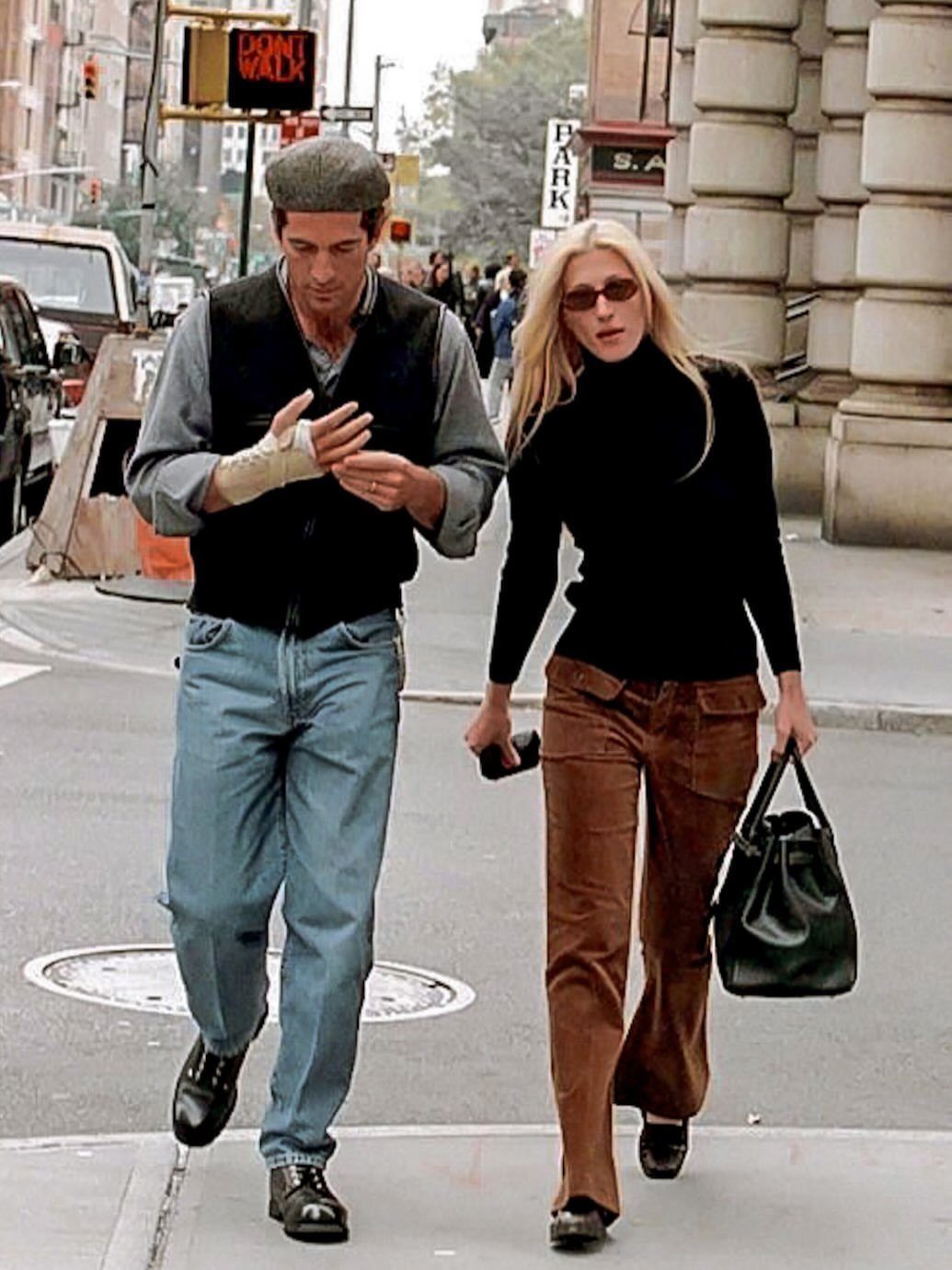 13 Carolyn Bessette Outfits You Can Actually Wear Today 8 Woman in black turtleneck and brown trousers walking on a city street with a man.