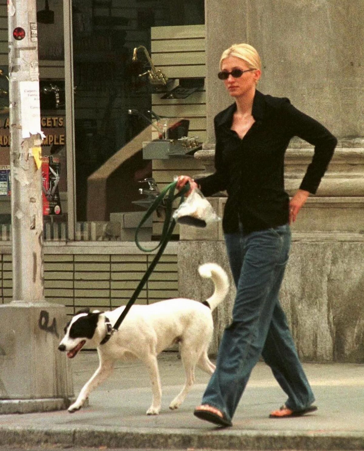13 Carolyn Bessette Outfits You Can Actually Wear Today 4 Woman in black button-down shirt and blue jeans walking a white dog.