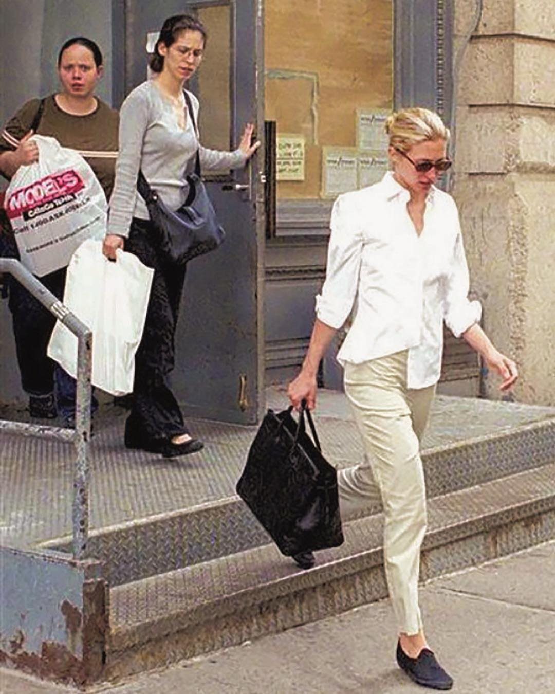 13 Carolyn Bessette Outfits You Can Actually Wear Today 1 Woman in oversized white shirt and beige pants carrying a large black leather bag.