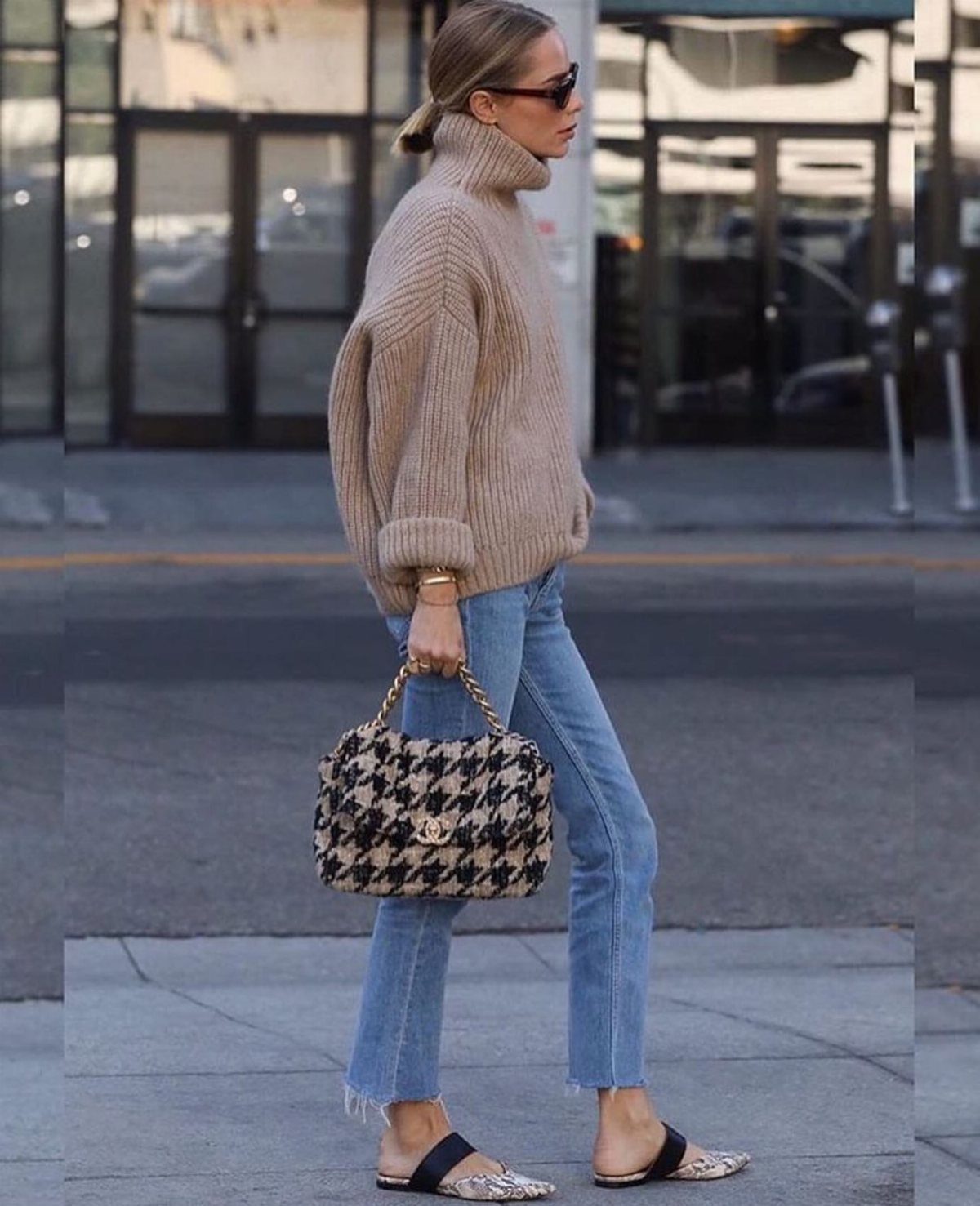 13 Carolyn Bessette Outfits You Can Actually Wear Today 10 Woman in beige oversized turtleneck sweater and light blue jeans carrying houndstooth handbag.