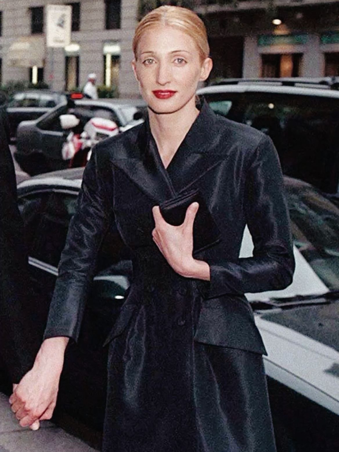 13 Carolyn Bessette Outfits You Can Actually Wear Today 11 Woman in a structured black satin coat holding a small black clutch bag.