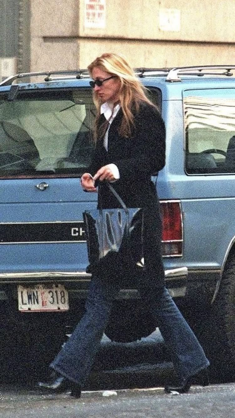 13 Carolyn Bessette Outfits You Can Actually Wear Today 13 Woman wearing a long black structured coat, white shirt, and flared blue jeans.