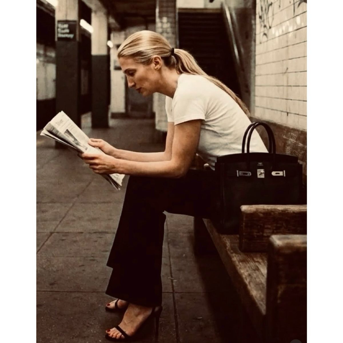 13 Carolyn Bessette Outfits You Can Actually Wear Today 7 Woman in white t-shirt and black trousers reading a newspaper in a subway.