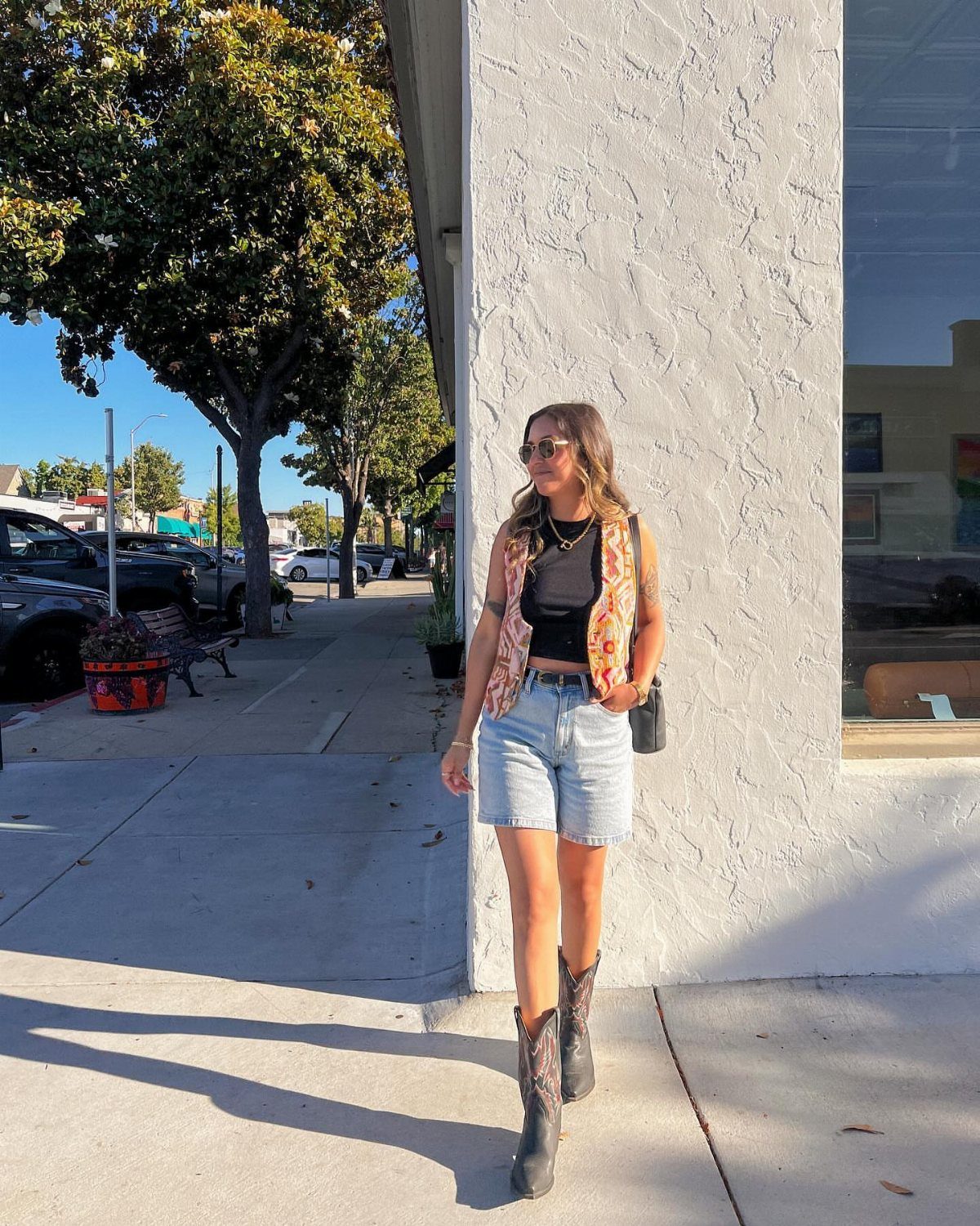 14 Coastal Cowgirl Outfits for Breezy Western Beach Style 14 Woman wearing a bohemian vest, denim shorts, and cowboy boots on a sidewalk.