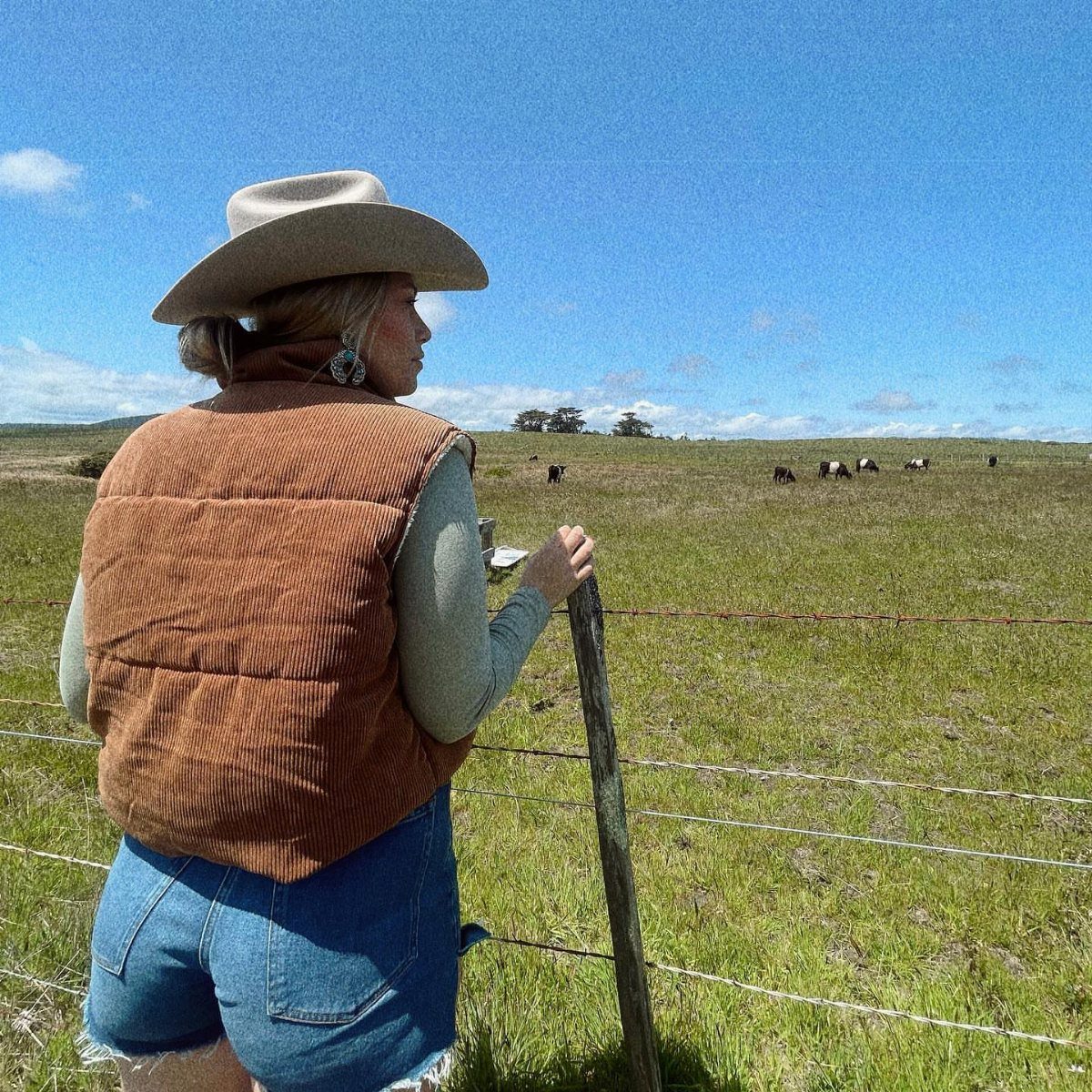 14 Coastal Cowgirl Outfits for Breezy Western Beach Style 11 Woman in cowboy hat and denim shorts standing in a grassy field with cows.