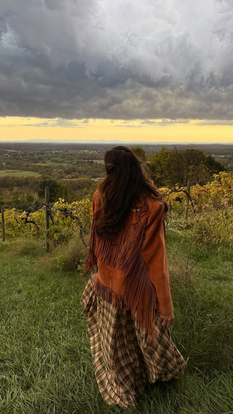 14 Coastal Cowgirl Outfits for Breezy Western Beach Style 2 Woman from behind wearing a brown fringe jacket and plaid skirt in a field.