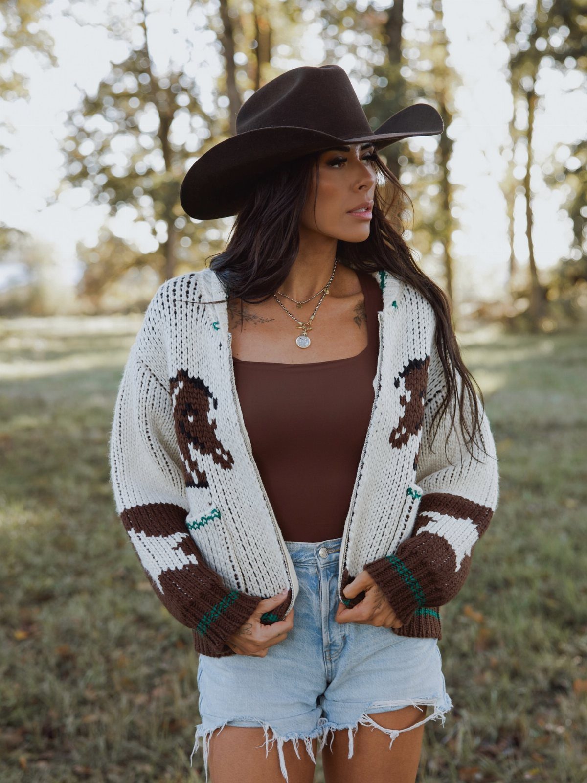 14 Coastal Cowgirl Outfits for Breezy Western Beach Style 8 Woman in brown cowboy hat and patterned cardigan wearing distressed denim shorts outdoors.