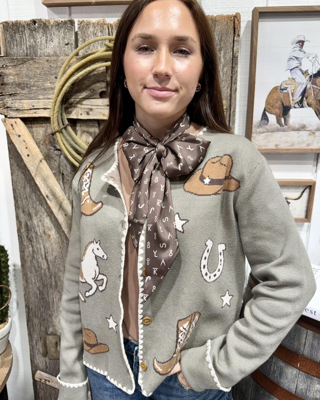 14 Coastal Cowgirl Outfits for Breezy Western Beach Style 9 Woman in a grey western-themed patched cardigan and brown neck scarf posing indoors.