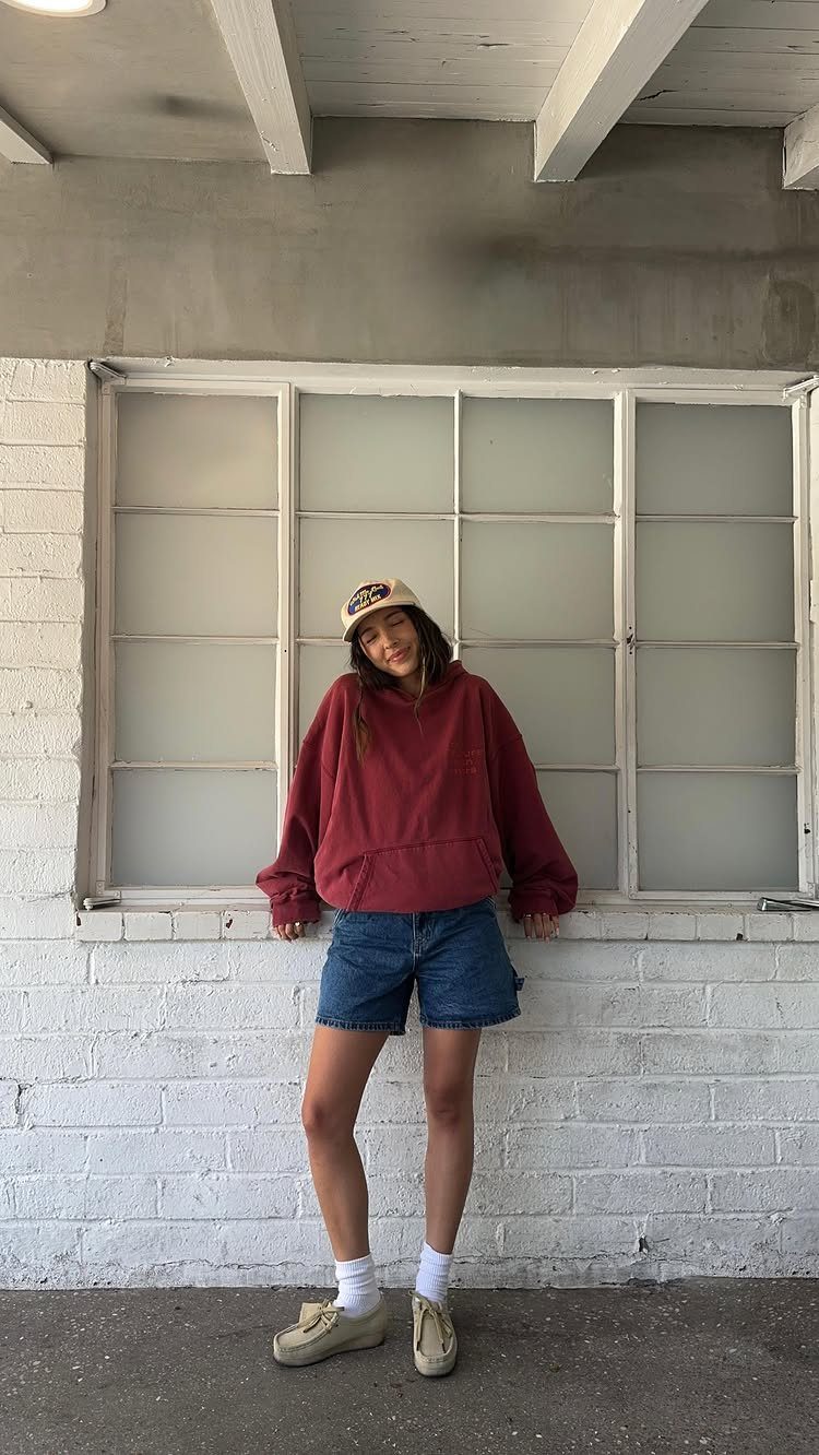 11 Comfortable Concert Outfits You'll Want to Wear in 2026 1 Person wearing a maroon hoodie and blue denim shorts standing by a window.