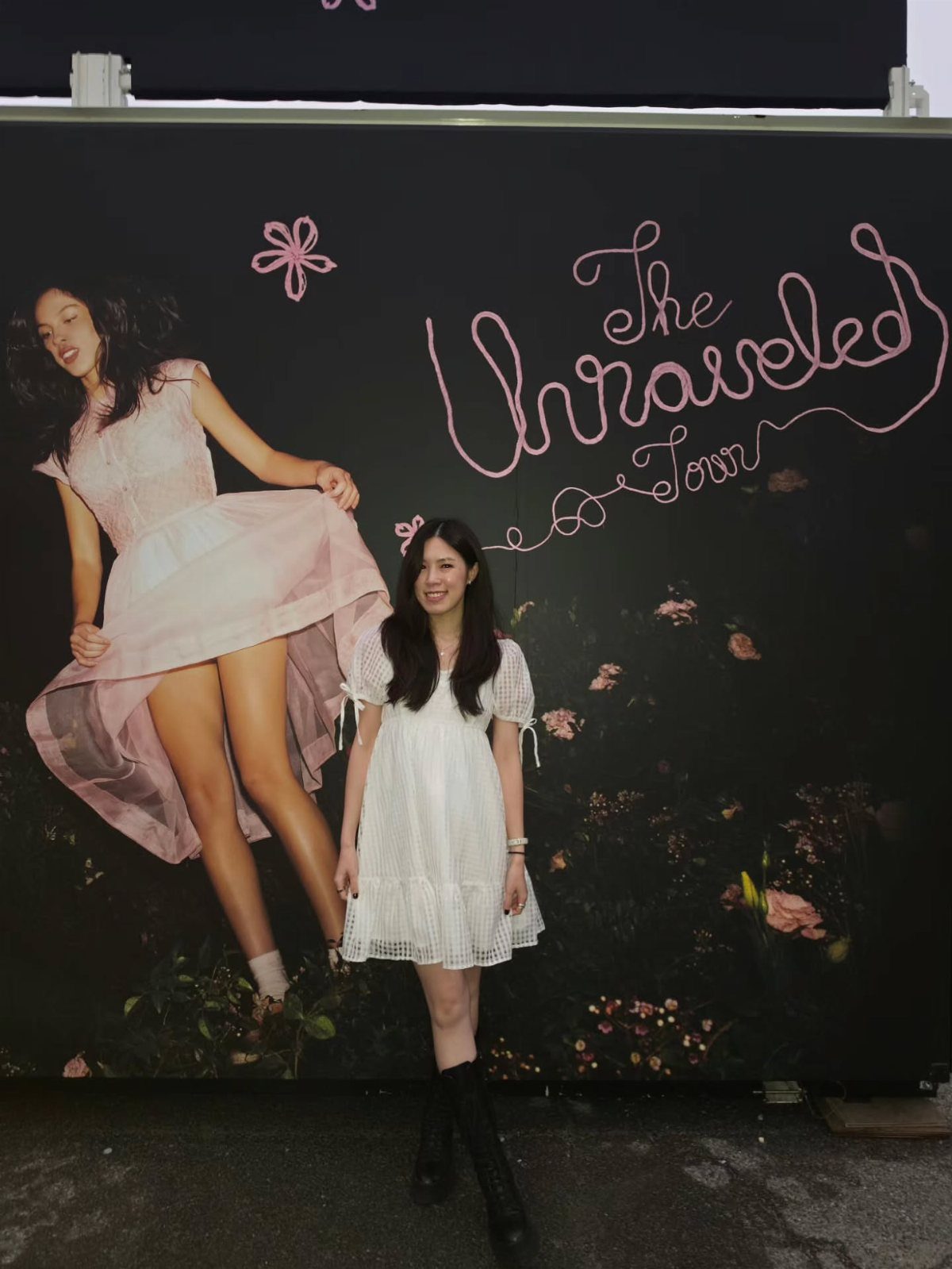 11 Comfortable Concert Outfits You'll Want to Wear in 2026 7 Woman in a short white flowy dress and black boots posing by tour backdrop.
