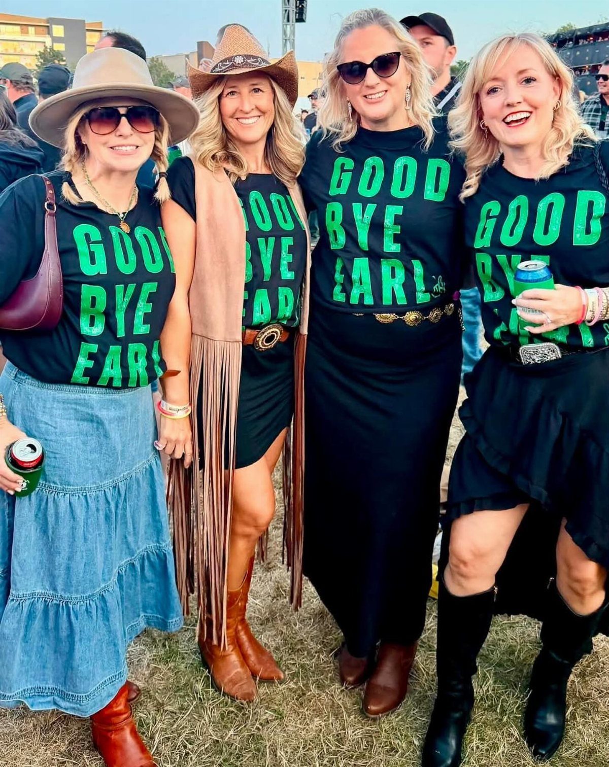 11 Comfortable Concert Outfits You'll Want to Wear in 2026 4 Four women wearing matching black graphic t-shirts and boots at an outdoor event.