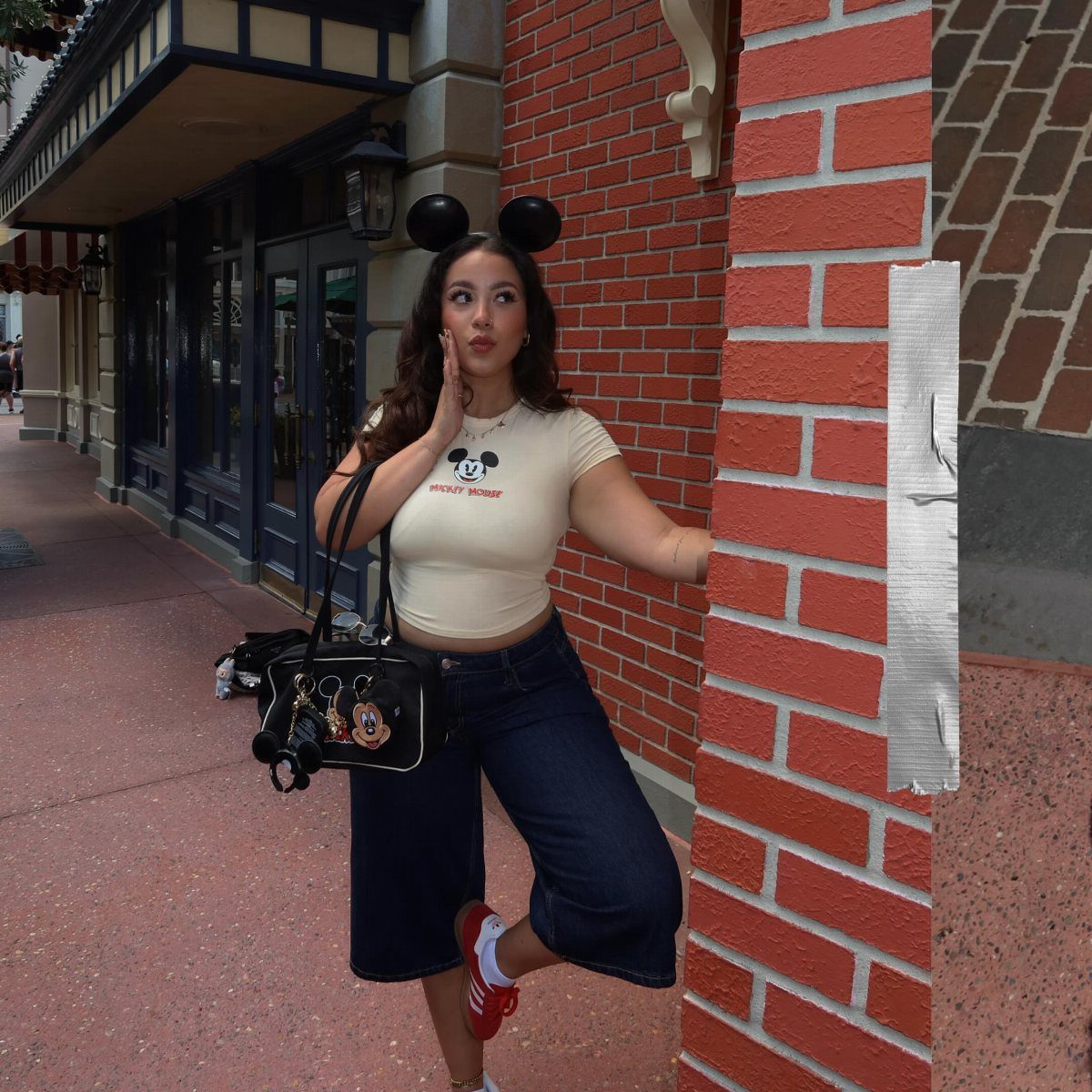 10 Cute Disney Outfits to Wear at the Parks in 2026 3 Woman in Mickey Mouse graphic tee and wide-leg cropped pants posing by brick wall.