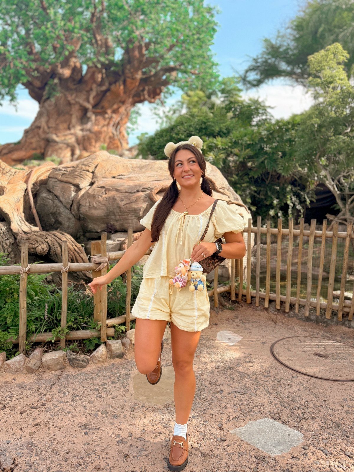 10 Cute Disney Outfits to Wear at the Parks in 2026 5 Woman in a yellow romper and Mickey ear headband posing by a large tree.
