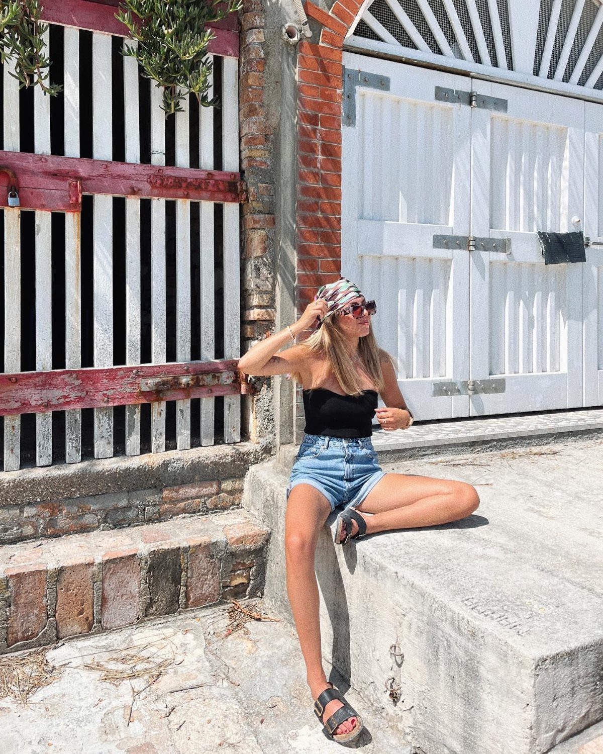 12 Cute Summer Outfits to Keep You Cool and Stylish 5 Woman wearing a black tank top and denim shorts sitting by a white door.