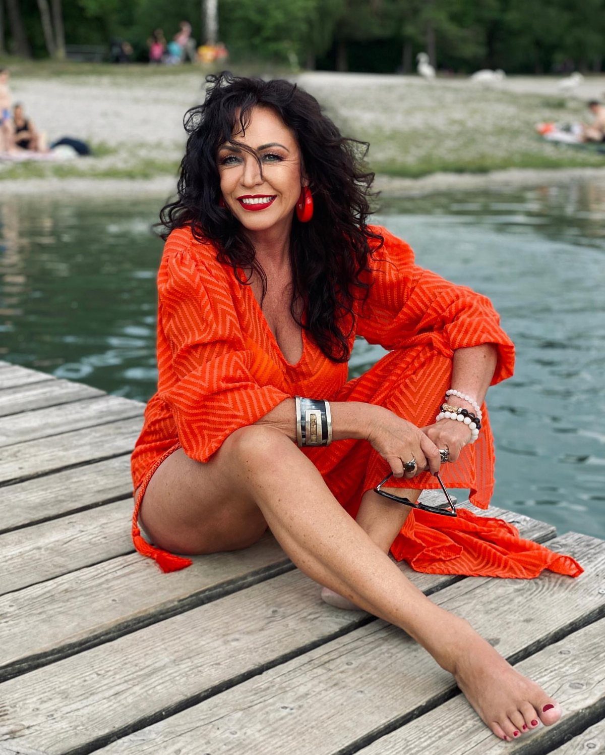 12 Cute Summer Outfits to Keep You Cool and Stylish 6 Woman in orange striped off-the-shoulder cover-up sitting on a wooden lake pier.