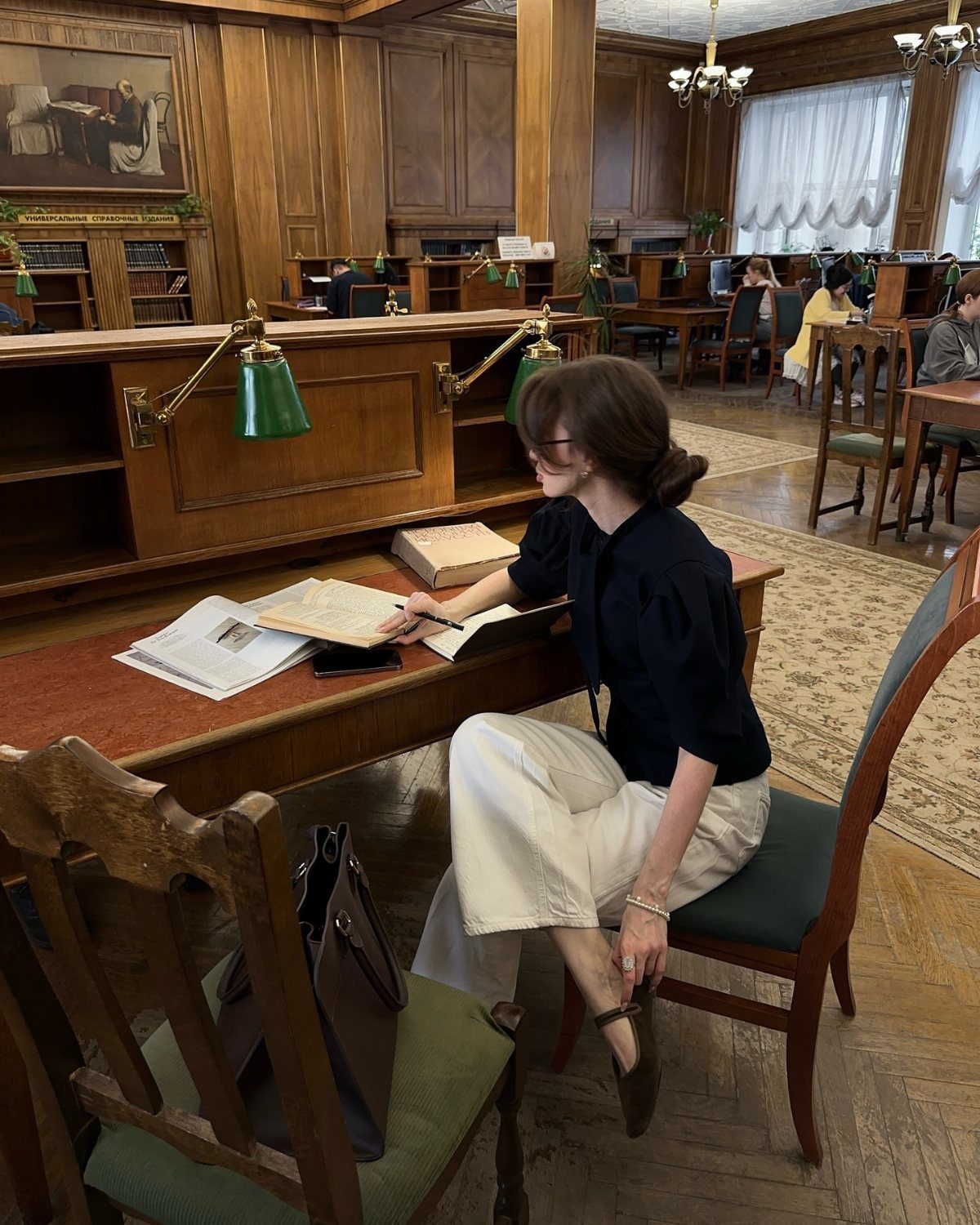 Dark Academia Outfits Female: 12 Ideas for Timeless Style 12 Woman in black blouse and wide-leg cream trousers studying at a wooden library desk.