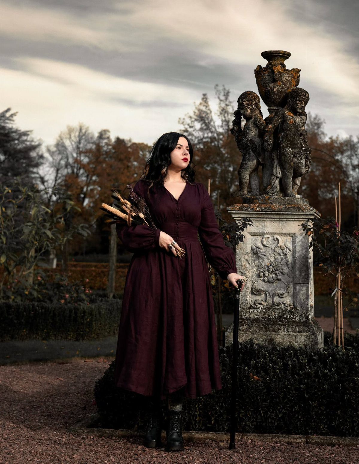 Dark Cottagecore Outfits: 8 Ideas from Dresses to Footwear 3 Woman in a deep burgundy vintage dress standing beside a stone cherub statue.