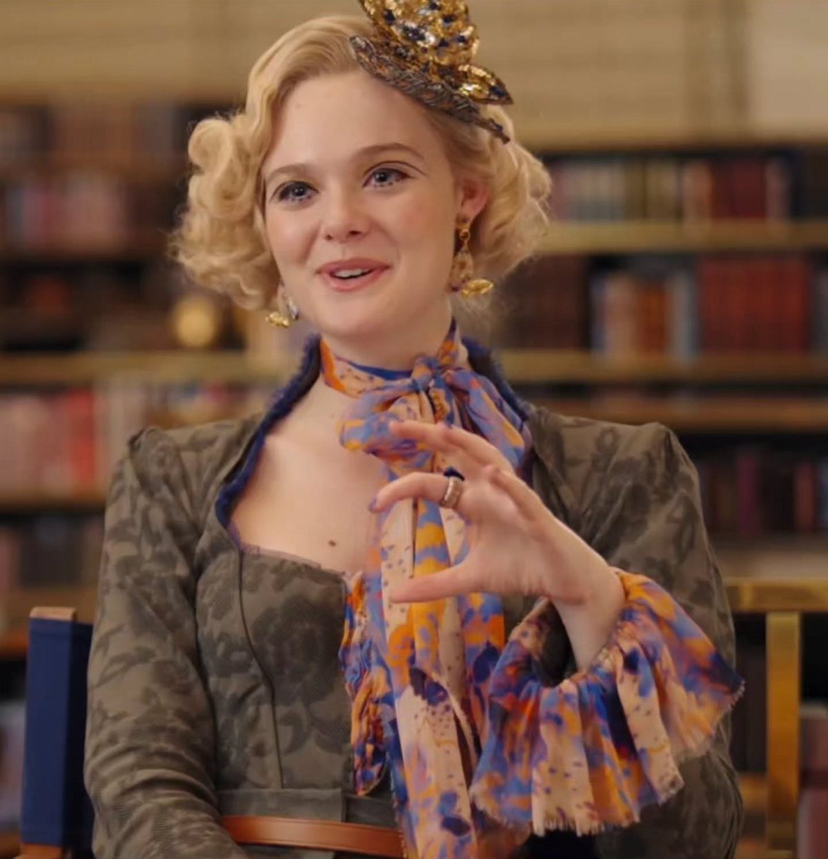 Effie Trinket Outfits: 10 Iconic Looks to Recreate 1 Woman with blonde curls wearing a small ornate gold fascinator hat and colorful scarf.