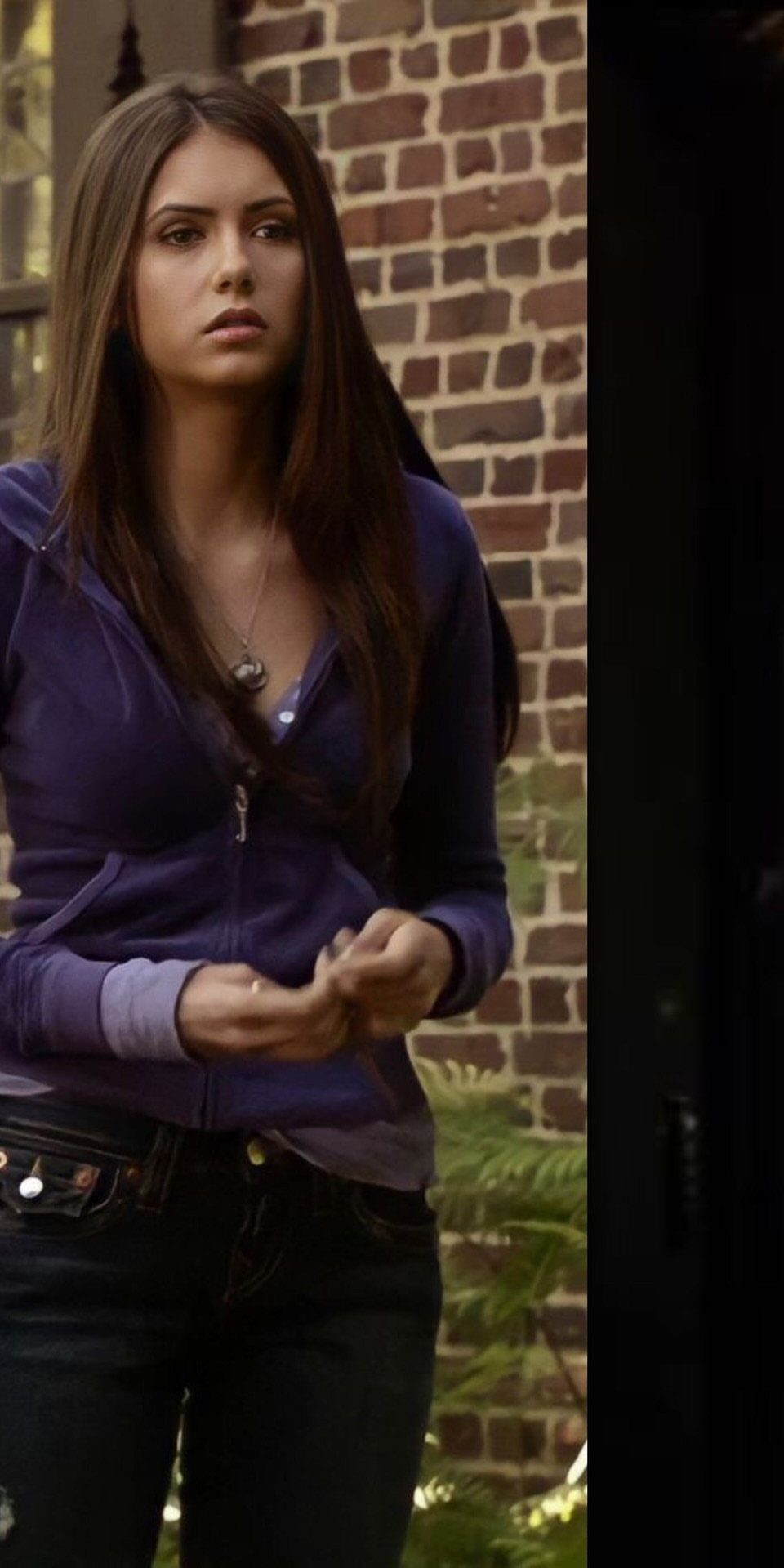 14 Elena Gilbert Outfits: Her Best Looks from the Show 7 Woman wearing a purple zip-up hoodie and dark jeans against a brick wall.